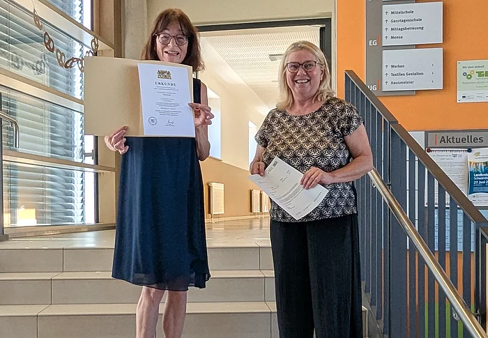 Zwei Frauen stehen lächelnd auf einer Treppe im Eingangsbereich der Telemann-Grundschule. Petra Koch hält ihre Pensionsurkunde in den Händen, daneben steht Schulleiterin Sabine Kunz mit weiteren Unterlagen.
