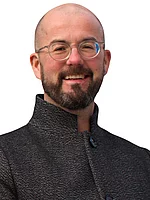 Ein Mann mit Glatze, Brille und Bart trägt eine hochgeschlossene Jacke und lächelt freundlich.