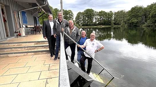 Fünf Männer stehen gemeinsam an einer Treppe, die von einer Terrasse direkt in einen See führt. Im Vordergrund verläuft ein Metallgeländer entlang der Treppe. Zwei Männer stehen auf den Stufen nahe am Wasser und halten lange Metallstangen, während die anderen daneben stehen und in die Kamera blicken. Links befindet sich ein Gebäude mit Terrasse und Sitzgelegenheiten, rechts erstreckt sich der ruhige See mit bewaldetem Ufer im Hintergrund. Der Himmel ist bewölkt. Die Aufnahme vermittelt den Eindruck eines gemeinsamen Termins oder einer Besichtigung an der Wasserwacht-Wachstation.