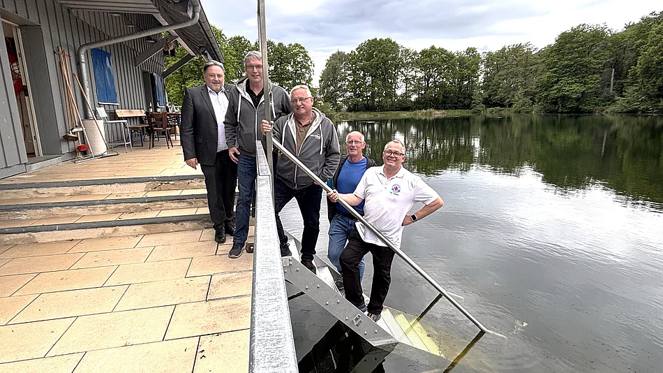 Fünf Männer stehen gemeinsam an einer Treppe, die von einer Terrasse direkt in einen See führt. Im Vordergrund verläuft ein Metallgeländer entlang der Treppe. Zwei Männer stehen auf den Stufen nahe am Wasser und halten lange Metallstangen, während die anderen daneben stehen und in die Kamera blicken. Links befindet sich ein Gebäude mit Terrasse und Sitzgelegenheiten, rechts erstreckt sich der ruhige See mit bewaldetem Ufer im Hintergrund. Der Himmel ist bewölkt. Die Aufnahme vermittelt den Eindruck eines gemeinsamen Termins oder einer Besichtigung an der Wasserwacht-Wachstation.