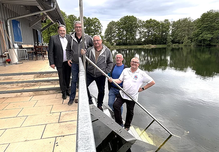Fünf Männer stehen gemeinsam an einer Treppe, die von einer Terrasse direkt in einen See führt. Im Vordergrund verläuft ein Metallgeländer entlang der Treppe. Zwei Männer stehen auf den Stufen nahe am Wasser und halten lange Metallstangen, während die anderen daneben stehen und in die Kamera blicken. Links befindet sich ein Gebäude mit Terrasse und Sitzgelegenheiten, rechts erstreckt sich der ruhige See mit bewaldetem Ufer im Hintergrund. Der Himmel ist bewölkt. Die Aufnahme vermittelt den Eindruck eines gemeinsamen Termins oder einer Besichtigung an der Wasserwacht-Wachstation.