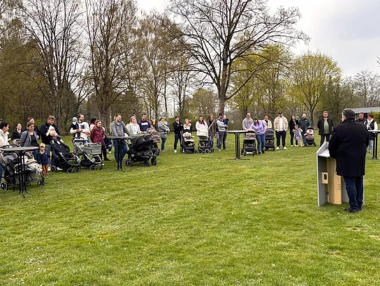 Mehrere Familien mit Kinderwagen stehen in einem Halbkreis auf einer Wiese und verfolgen eine Ansprache bei der Baumpflanzaktion.