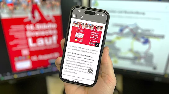 Eine Hand hält ein Smartphone vor zwei unscharf dargestellten Computerbildschirmen. Auf dem Smartphone ist die mobile Ansicht einer Website zum „16. Städtedreieckslauf“ zu sehen, darunter Informationen zur Veranstaltung. Auf dem linken Bildschirm im Hintergrund ist ein rotes Veranstaltungsplakat erkennbar, rechts eine Website mit einer Streckenübersicht oder Grafik. Die Aufnahme vermittelt den Eindruck einer modernen, digitalen Informationsplattform für den Städtedreieckslauf.