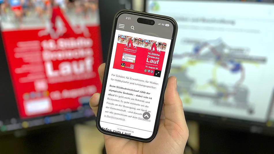 Eine Hand hält ein Smartphone vor zwei unscharf dargestellten Computerbildschirmen. Auf dem Smartphone ist die mobile Ansicht einer Website zum „16. Städtedreieckslauf“ zu sehen, darunter Informationen zur Veranstaltung. Auf dem linken Bildschirm im Hintergrund ist ein rotes Veranstaltungsplakat erkennbar, rechts eine Website mit einer Streckenübersicht oder Grafik. Die Aufnahme vermittelt den Eindruck einer modernen, digitalen Informationsplattform für den Städtedreieckslauf.