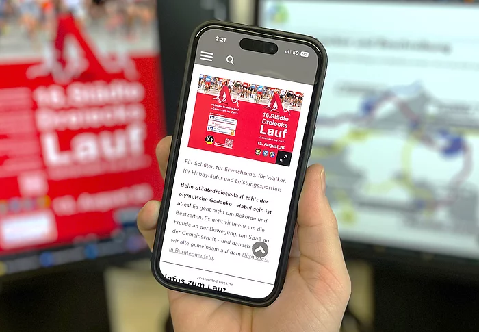Eine Hand hält ein Smartphone vor zwei unscharf dargestellten Computerbildschirmen. Auf dem Smartphone ist die mobile Ansicht einer Website zum „16. Städtedreieckslauf“ zu sehen, darunter Informationen zur Veranstaltung. Auf dem linken Bildschirm im Hintergrund ist ein rotes Veranstaltungsplakat erkennbar, rechts eine Website mit einer Streckenübersicht oder Grafik. Die Aufnahme vermittelt den Eindruck einer modernen, digitalen Informationsplattform für den Städtedreieckslauf.