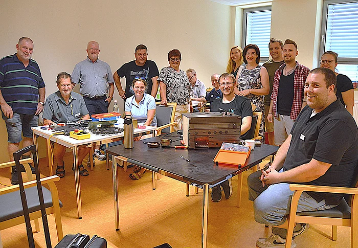Eine Gruppe von rund einem Dutzend Personen steht und sitzt in einem hellen Raum um mehrere Tische, auf denen verschiedene Elektrogeräte, Werkzeugkästen, Messgeräte und Kleinteile liegen. Im Mittelpunkt steht ein geöffnetes Hi-Fi-Gerät, an dem gearbeitet wird. Die Teilnehmenden lächeln in die Kamera und vermitteln den Eindruck einer gemeinschaftlichen und engagierten Atmosphäre. Das Foto entstand anlässlich des ersten Geburtstags des Reparatur-Cafés.