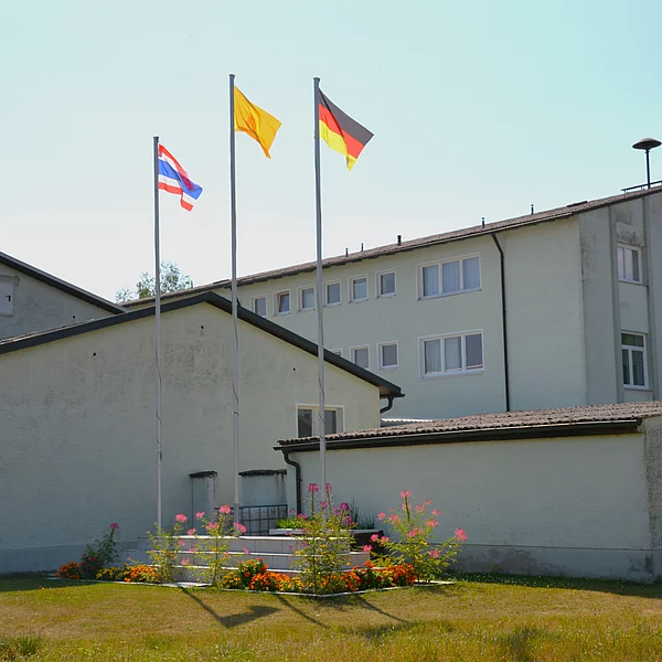 Ein großer Gebäudekomplex umgeben von Rasen. Im Vordergrund drei Fahnenmasten mit der Thailänischen, Deutschen und Buddhistischen Flagge.