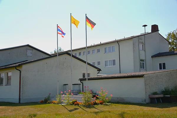 Ein großer Gebäudekomplex umgeben von Rasen. Im Vordergrund drei Fahnenmasten mit der Thailänischen, Deutschen und Buddhistischen Flagge.