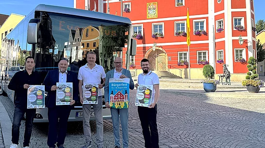 Vor dem historischen Rathaus in Burglengenfeld steht der Event-Bus des Städtedreiecks. Fünf Vertreter der Stadt und des Organisationsteams präsentieren die Programmflyer für das Bürgerfest sowie das Veranstaltungsplakat und werben gemeinsam für das Festwochenende.
