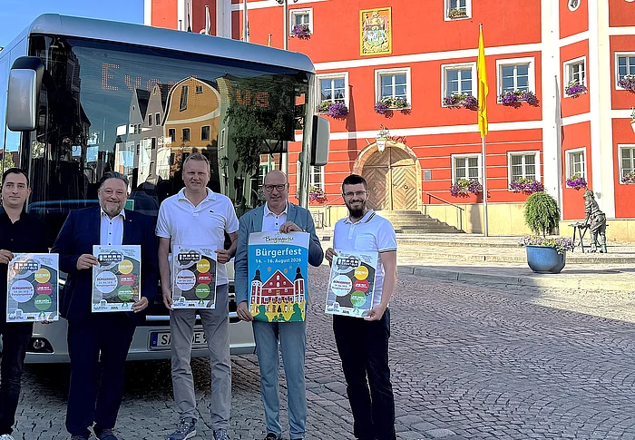 Vor dem historischen Rathaus in Burglengenfeld steht der Event-Bus des Städtedreiecks. Fünf Vertreter der Stadt und des Organisationsteams präsentieren die Programmflyer für das Bürgerfest sowie das Veranstaltungsplakat und werben gemeinsam für das Festwochenende.