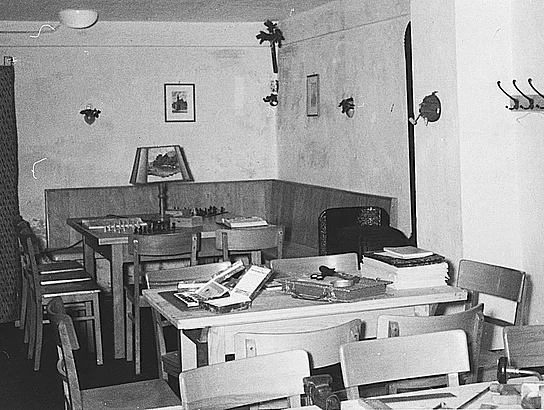 Ein historisches Schwarz-Weiß-Foto zeigt einen Aufenthaltsraum des Jugendheims. Mehrere schlichte Holztische und Stühle sind aufgestellt, auf einem Tisch liegen ein Schachspiel und weitere Gesellschaftsspiele. Die Einrichtung wirkt einfach, aber gemütlich.