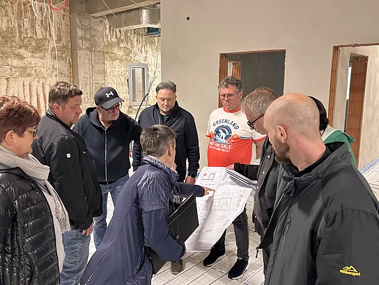 Eine Gruppe von Personen steht in einem unfertigen Innenraum eines Gebäudes und bespricht Baupläne zwischen freiliegenden Wänden und Installationen.