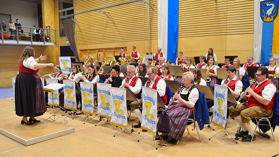 Eine große Blaskapelle in Tracht spielt auf einer Bühne, während die Dirigentin vor dem Orchester steht und die Musikerinnen und Musiker anleitet.