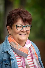 Eine ältere Frau mit kurzen Haaren, Brille und buntem Schal lächelt freundlich.