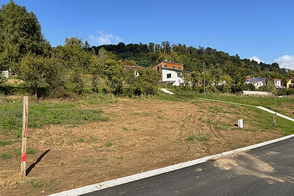 Ein leicht ansteigendes, unbebautes Grundstück mit brauner Erde liegt an einer neu asphaltierten Straße; im Hintergrund stehen einzelne Häuser am Waldrand unter blauem Himmel.