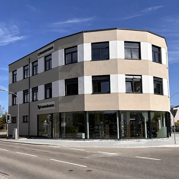 Außenansicht der neuen Stadtbücherei Teublitz an einer Straßenecke bei sonnigem Wetter. Das moderne, mehrgeschossige Gebäude mit großen Glasfronten im Erdgeschoss steht an einer Kreuzung. Durch die Fenster ist der helle Innenbereich der Bücherei mit Regalen sichtbar. Über dem Eingang sowie an der Glasfassade ist die Beschriftung „Stadtbücherei“ angebracht. Im Vordergrund verläuft die Straße, links rahmt ein Baum das Bild ein.
