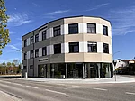 Außenansicht der neuen Stadtbücherei Teublitz an einer Straßenecke bei sonnigem Wetter. Das moderne, mehrgeschossige Gebäude mit großen Glasfronten im Erdgeschoss steht an einer Kreuzung. Durch die Fenster ist der helle Innenbereich der Bücherei mit Regalen sichtbar. Über dem Eingang sowie an der Glasfassade ist die Beschriftung „Stadtbücherei“ angebracht. Im Vordergrund verläuft die Straße, links rahmt ein Baum das Bild ein.