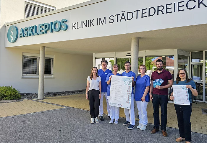 Sieben Personen stehen vor dem Eingang der Asklepios Klinik im Städtedreieck, darunter mehrere Mitarbeitende in medizinischer Berufskleidung; zwei von ihnen halten Plakate zur Veranstaltungsreihe „Klinik im Dialog 2026“ in die Kamera.