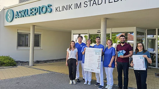 Sieben Personen stehen vor dem Eingang der Asklepios Klinik im Städtedreieck, darunter mehrere Mitarbeitende in medizinischer Berufskleidung; zwei von ihnen halten Plakate zur Veranstaltungsreihe „Klinik im Dialog 2026“ in die Kamera.