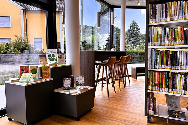Ein hoher Lerntisch mit Barhockern steht direkt an einer großen Glasfront mit Blick ins Grüne. Daneben präsentieren kleine Ausstellungsflächen aktuelle Buchempfehlungen. Helle Holzböden und moderne Regale sorgen für eine freundliche Lernumgebung.