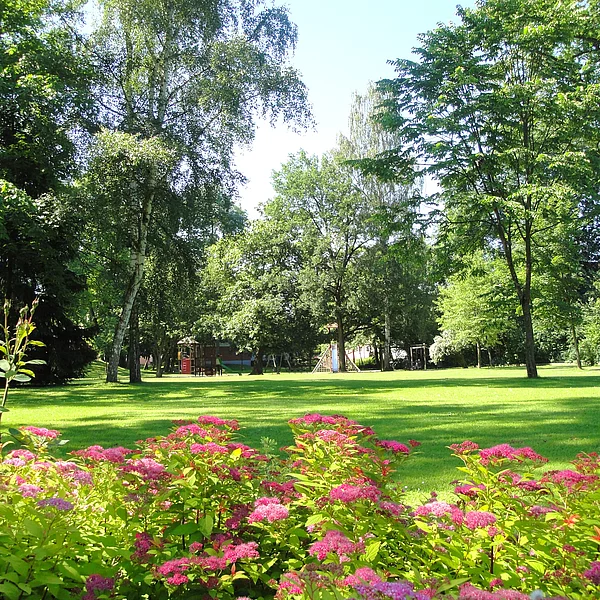 Blühende Sträucher vor einer saftig grünen Liegewiese. Im Hintergrund unzählige Bäume und ein großer Spielplatz.