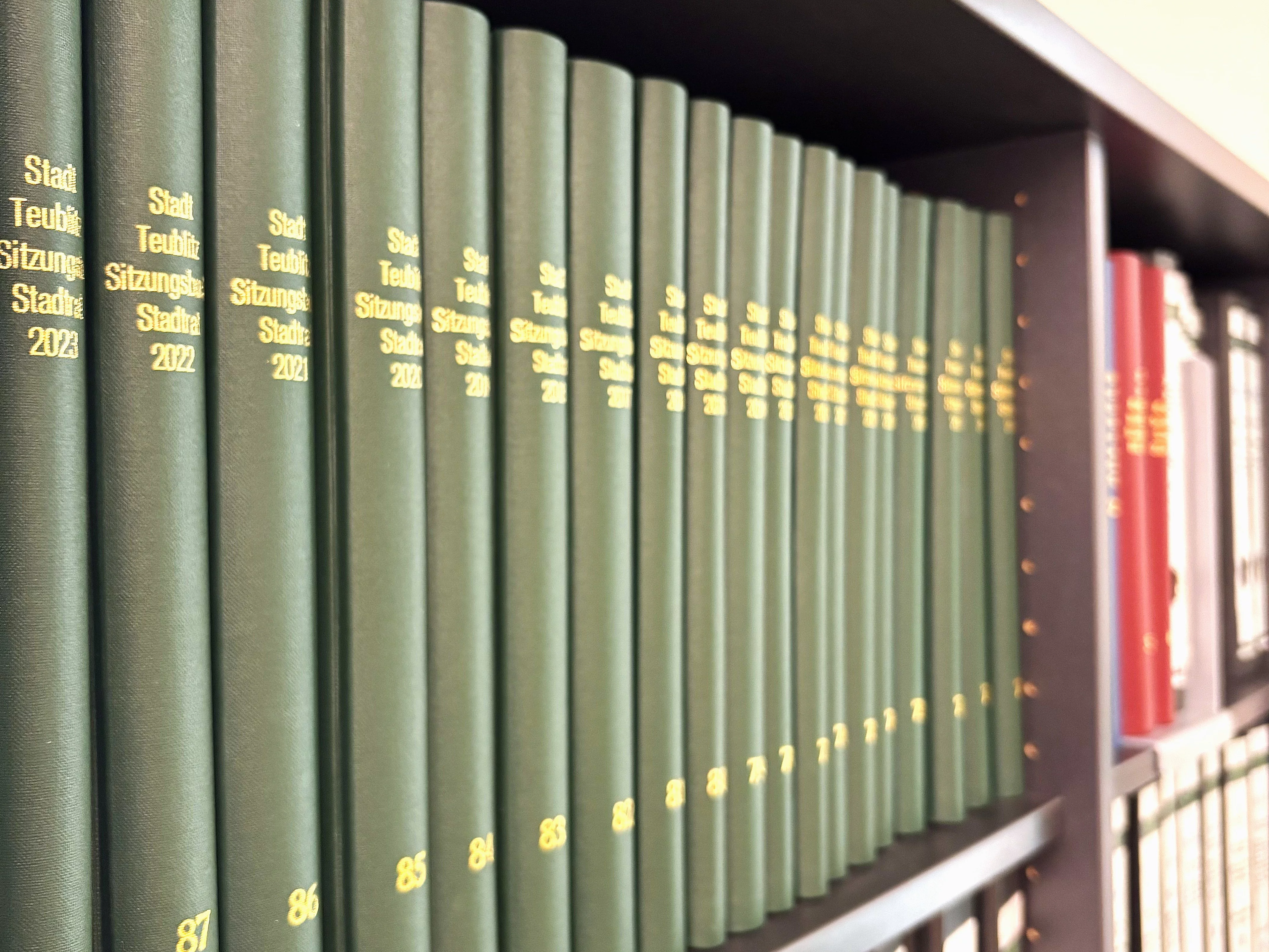 Ein Regal zeigt eine Reihe grüner gebundener Bücher mit goldener Beschriftung zu Stadtratssitzungen, darunter Ordner im unteren Fach.