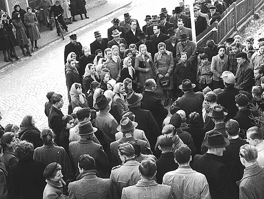 Eine historische Schwarz-Weiß-Aufnahme aus erhöhter Perspektive zeigt eine große Menschenmenge vor dem Jugendheim. Erwachsene, Jugendliche und Kinder bilden einen Kreis um mehrere Redner. Die Straße und die umliegenden Gebäude sind dicht mit Besucherinnen und Besuchern gefüllt.