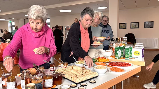 Mehrere Seniorinnen bedienen sich an einem reich gedeckten Frühstücksbuffet im Mehrgenerationenhaus Teublitz. Auf den Tischen stehen unter anderem verschiedene Aufstriche, Brot und Gebäck, Cracker, Butter sowie frisches Gemüse. Im Hintergrund sitzen weitere Gäste gemeinsam an langen Tischen und unterhalten sich.