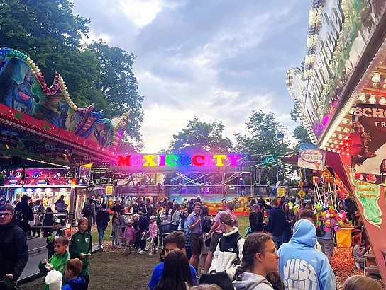 Besucher drängen sich auf einem Volksfestplatz zwischen Ständen und Fahrgeschäften, während in der Mitte ein bunt beleuchtetes Fahrgeschäft mit der Aufschrift „Mexico City“ zu sehen ist.