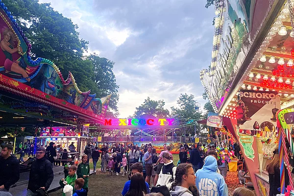 Besucher drängen sich auf einem Volksfestplatz zwischen Ständen und Fahrgeschäften, während in der Mitte ein bunt beleuchtetes Fahrgeschäft mit der Aufschrift „Mexico City“ zu sehen ist.