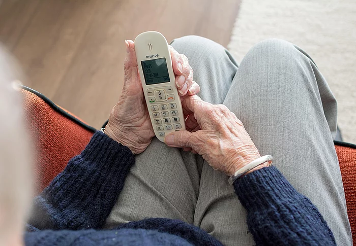 Eine ältere Person sitzt in einem Sessel und hält ein schnurloses Telefon in beiden Händen. Das Symbolbild begleitet die Ankündigung eines Vortrags zum Thema Schockanrufe und Telefonbetrug.