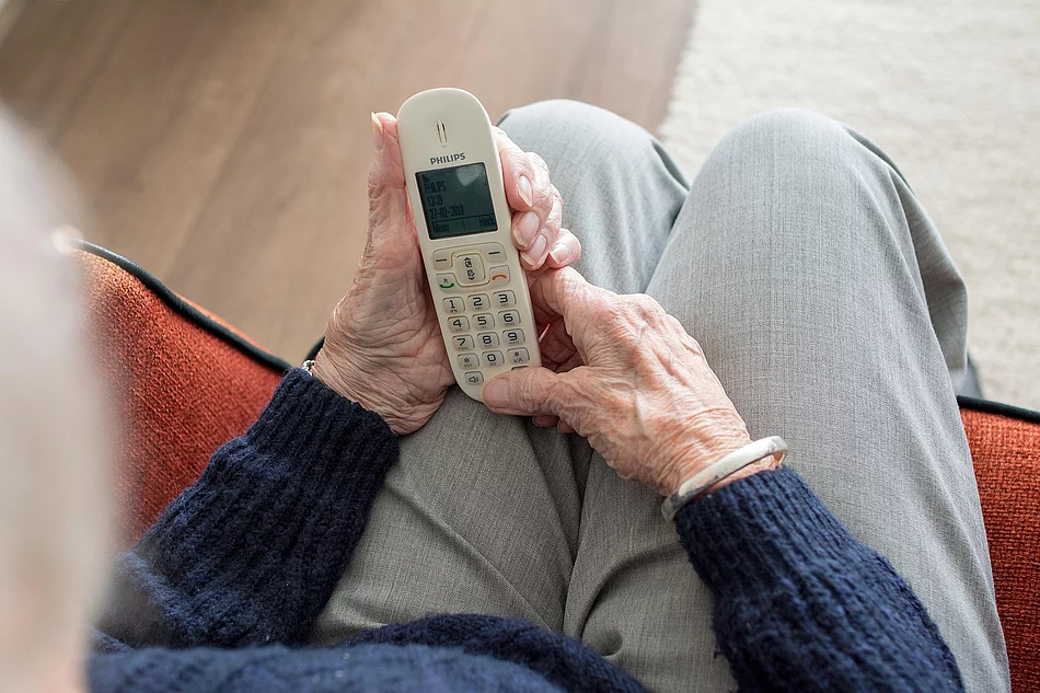 Eine ältere Person sitzt in einem Sessel und hält ein schnurloses Telefon in beiden Händen. Das Symbolbild begleitet die Ankündigung eines Vortrags zum Thema Schockanrufe und Telefonbetrug.