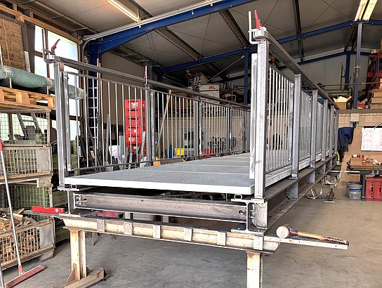 In einer Werkhalle steht eine neu gefertigte Metall-Fußgängerbrücke mit Geländer auf einem Arbeitstisch, umgeben von Werkzeugen und Baumaterialien.