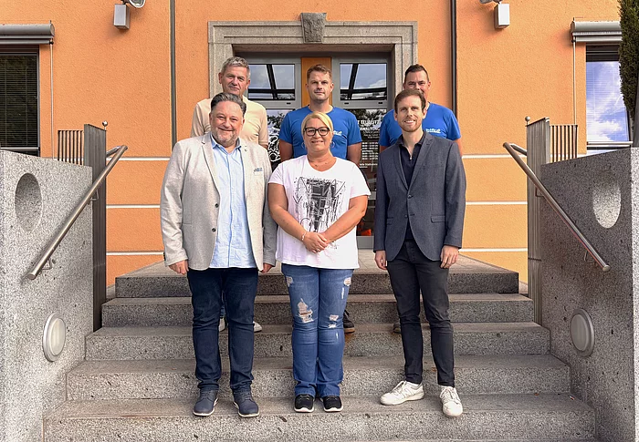 Sechs Personen stehen für ein Gruppenfoto auf der Treppe vor einem orangefarbenen Gebäude. Im Mittelpunkt steht Kathrin Steger. Neben ihr stehen zwei Männer, dahinter drei weitere Männer. Die Gruppe blickt lächelnd in die Kamera und heißt die neue Mitarbeiterin im Mensa-Team willkommen.