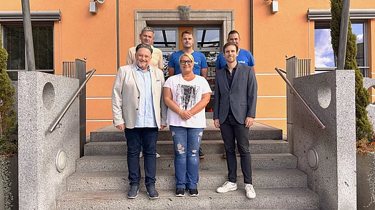 Sechs Personen stehen für ein Gruppenfoto auf der Treppe vor einem orangefarbenen Gebäude. Im Mittelpunkt steht Kathrin Steger. Neben ihr stehen zwei Männer, dahinter drei weitere Männer. Die Gruppe blickt lächelnd in die Kamera und heißt die neue Mitarbeiterin im Mensa-Team willkommen.
