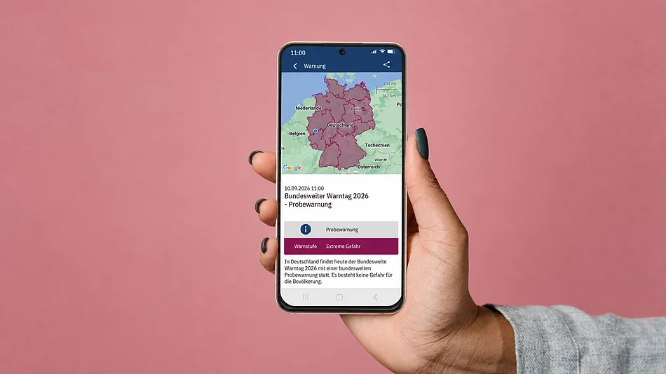 Symbolbild zum Bundesweiten Warntag 2026: Eine Hand hält ein Smartphone vor rosafarbenem Hintergrund, auf dessen Display eine Karte Deutschlands und eine Probewarnung zum Warntag am 10. September 2026 zu sehen sind.