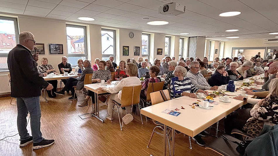 In einem hellen Veranstaltungsraum sitzen zahlreiche Seniorinnen und Senioren an gedeckten Tischen bei Kaffee und Kuchen. Im Vordergrund steht ein Mann mit Mikrofon und spricht zur Gruppe. Die Gäste hören aufmerksam zu, einige lachen oder unterhalten sich. Große Fenster, Bilder an den Wänden und eine freundliche Atmosphäre prägen den Raum.