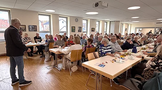 In einem hellen Veranstaltungsraum sitzen zahlreiche Seniorinnen und Senioren an gedeckten Tischen bei Kaffee und Kuchen. Im Vordergrund steht ein Mann mit Mikrofon und spricht zur Gruppe. Die Gäste hören aufmerksam zu, einige lachen oder unterhalten sich. Große Fenster, Bilder an den Wänden und eine freundliche Atmosphäre prägen den Raum.
