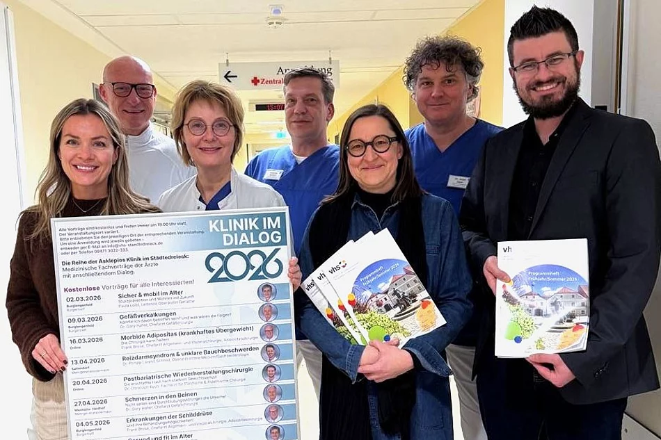 Mehrere Personen stehen gemeinsam auf einem Krankenhausflur, lächeln in die Kamera und präsentieren ein großes Veranstaltungsplakat sowie Programmhefte zu einer medizinischen Vortragsreihe.
