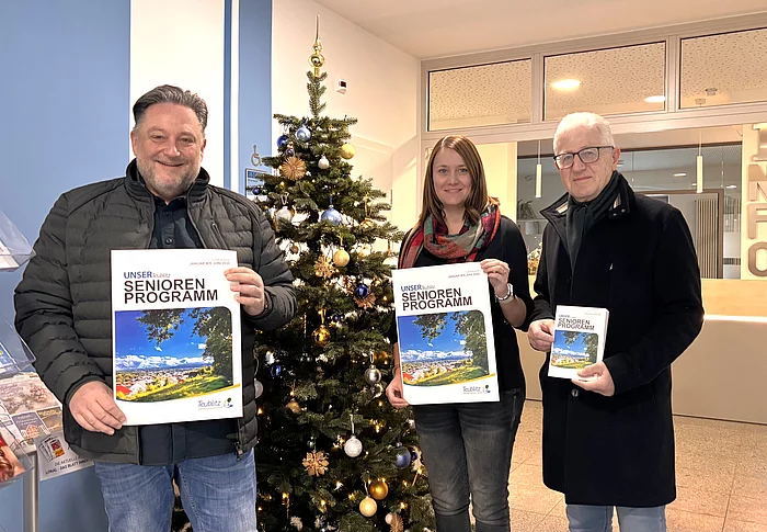 Drei Personen stehen in einem Eingangsbereich vor einem geschmückten Weihnachtsbaum und halten jeweils das neue Seniorenprogramm in der Hand. Die Hefte zeigen ein Titelbild mit Landschaftsfotografie. Alle drei lächeln in die Kamera, und im Hintergrund ist ein Infobereich des Gebäudes zu sehen.