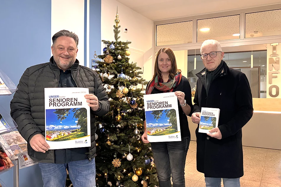 Drei Personen stehen in einem Eingangsbereich vor einem geschmückten Weihnachtsbaum und halten jeweils das neue Seniorenprogramm in der Hand. Die Hefte zeigen ein Titelbild mit Landschaftsfotografie. Alle drei lächeln in die Kamera, und im Hintergrund ist ein Infobereich des Gebäudes zu sehen.