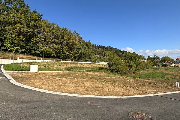 Ein leicht ansteigendes, unbebautes Grundstück mit brauner Erde liegt an einer neu asphaltierten Straße; im Hintergrund stehen einzelne Häuser am Waldrand unter blauem Himmel.