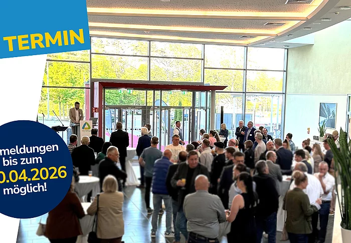 In einer großen, hellen Eingangshalle mit Glasfront stehen zahlreiche Menschen an Stehtischen und unterhalten sich, während links ein Redner an einem Pult spricht und darüber ein grafischer Hinweis mit Anmeldedatum eingeblendet ist.