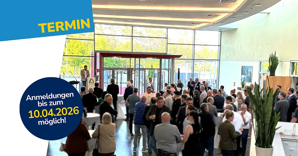 In einer großen, hellen Eingangshalle mit Glasfront stehen zahlreiche Menschen an Stehtischen und unterhalten sich, während links ein Redner an einem Pult spricht und darüber ein grafischer Hinweis mit Anmeldedatum eingeblendet ist.