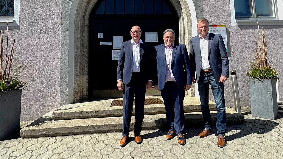 Drei Männer stehen bei Sonnenschein vor dem Eingang eines Rathauses und posieren für ein Foto.