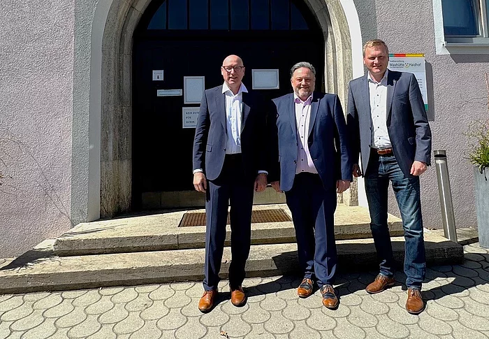 Drei Männer stehen bei Sonnenschein vor dem Eingang eines Rathauses und posieren für ein Foto.
