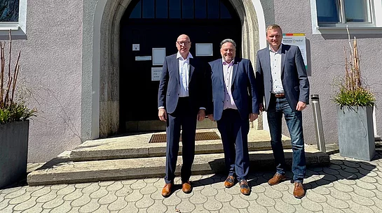 Drei Männer stehen bei Sonnenschein vor dem Eingang eines Rathauses und posieren für ein Foto.