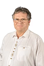 Ein älterer Mann mit Brille und grauem Haar trägt ein helles, traditionell wirkendes Hemd und schaut ruhig in die Kamera.