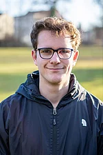 Ein junger Mann mit Brille und lockigem Haar steht im Freien und blickt freundlich in die Kamera.