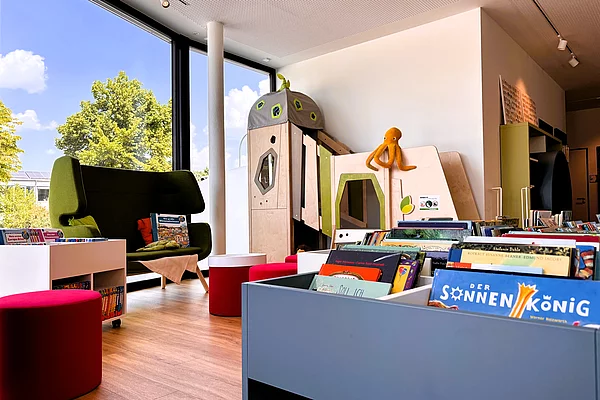 Ein modern gestalteter Kinderbereich mit niedrigen Bücherkisten, roten Sitzhockern und einer kleinen Spiel- beziehungsweise Rückzugsecke in Holzoptik. Eine Stoffkrake sitzt dekorativ auf dem Spielmöbel. Große Fenster sorgen für viel Tageslicht und eine helle Atmosphäre.